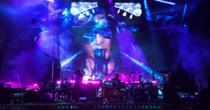 Front View of the Guns N Roses Drummer on Stage with Slashs Face Projected on the Back Screen in Blue and Purple Color Lighting My Dirty Life and Times