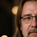 A close-up photo of a middle-aged person with long light brown hair, glasses, and a beard, shown from the nose up with only half of their face visible. The background is softly blurred with warm lights.